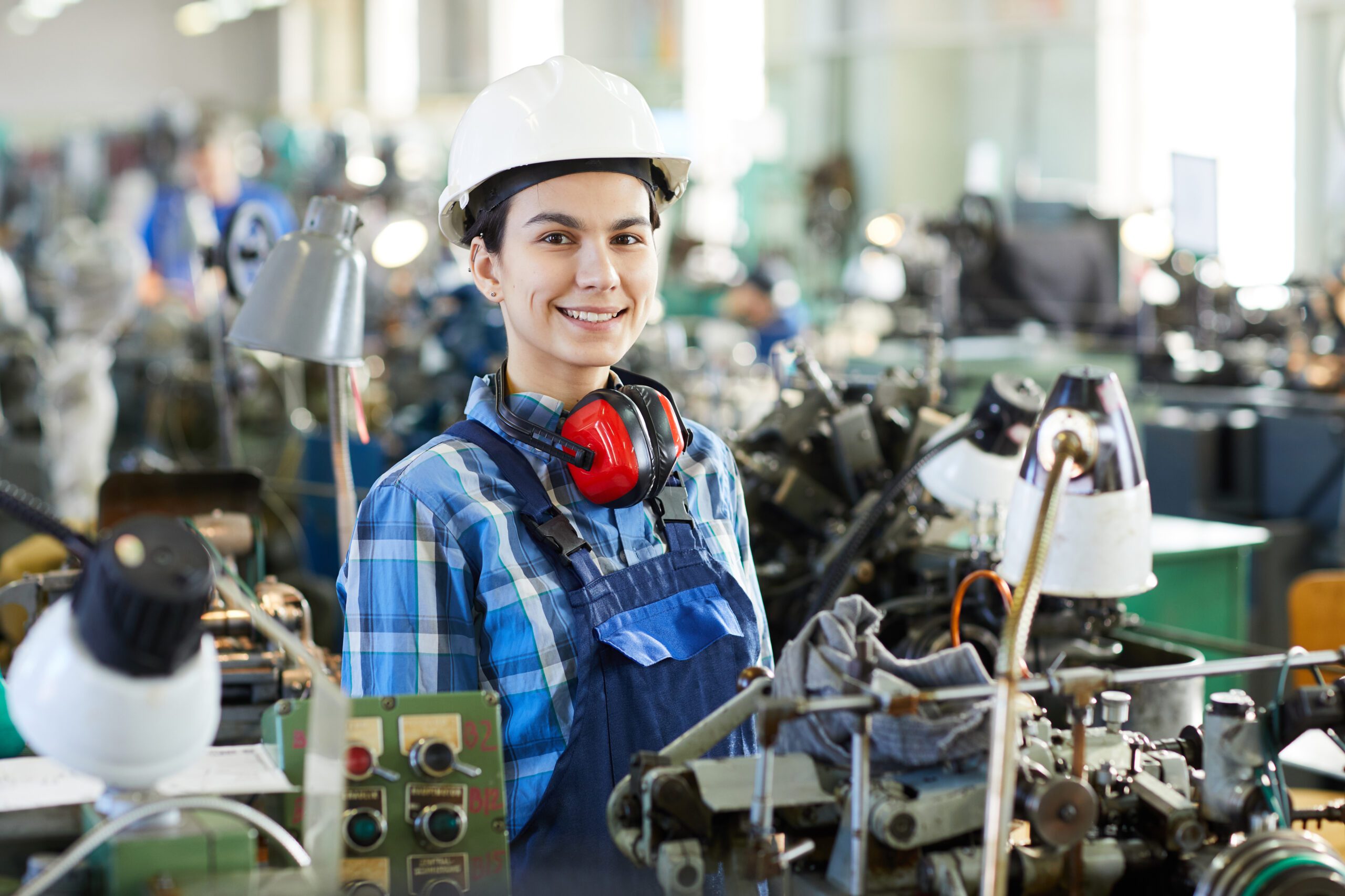 Positive confident attractive masculine lady engineer in hard hat standing at machine for producing clock movements and looking at camera, she working at manufacturing factory